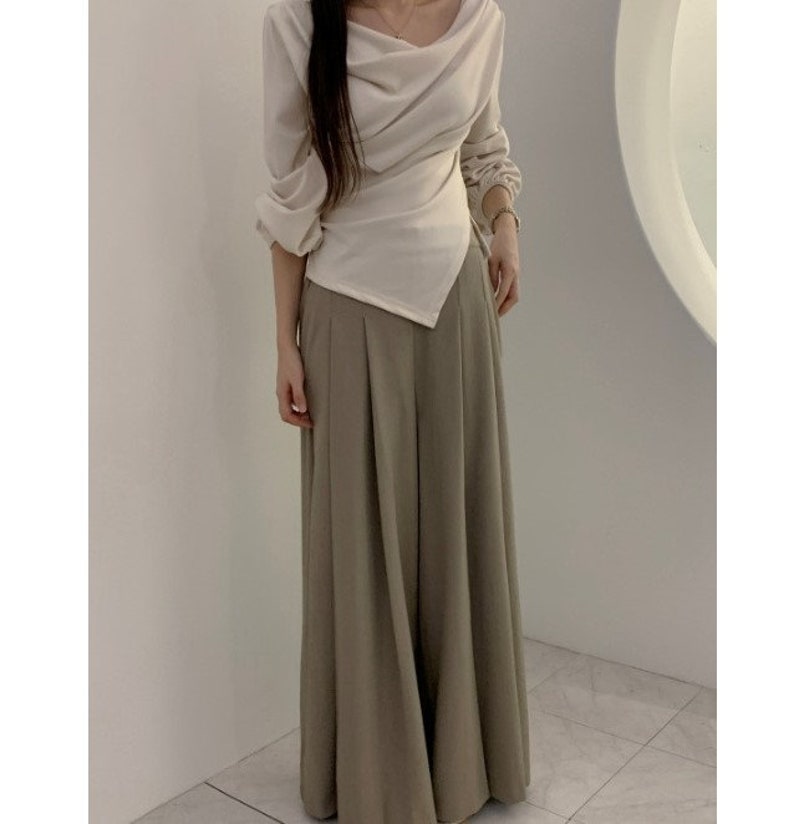 May include: A cream-colored long-sleeve top with an asymmetrical design and a draped neckline. Paired with wide-leg, pleated, olive-green pants. The outfit is a modern take on a classic style.