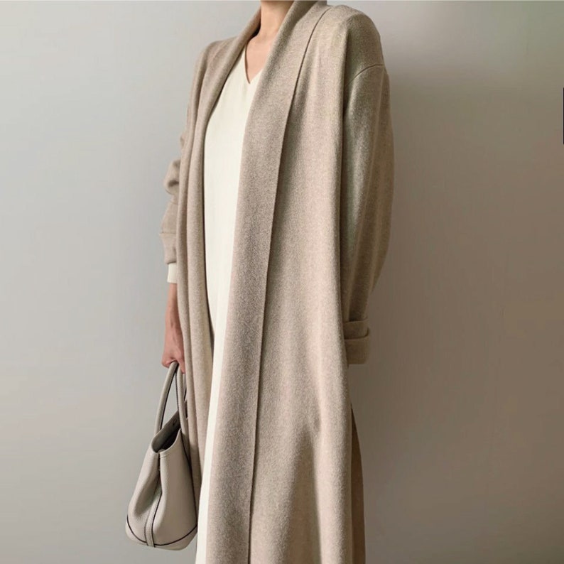May include: A long, beige cardigan with a shawl collar is worn over a white dress. The cardigan has long sleeves and a relaxed fit. A white handbag is held in the hand. The outfit is neutral-toned and minimalist.