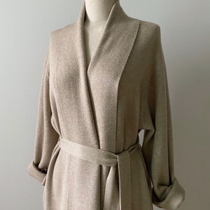 May include: A beige, long-sleeved robe with a shawl collar and a matching belt. The robe is made of a soft, textured fabric and has rolled-up sleeves. The robe is displayed on a mannequin.