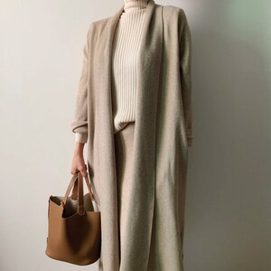 May include: A beige long cardigan and matching skirt are worn over a cream-colored ribbed turtleneck. A brown leather tote bag is held in the hand. The outfit is neutral-toned and minimalist.