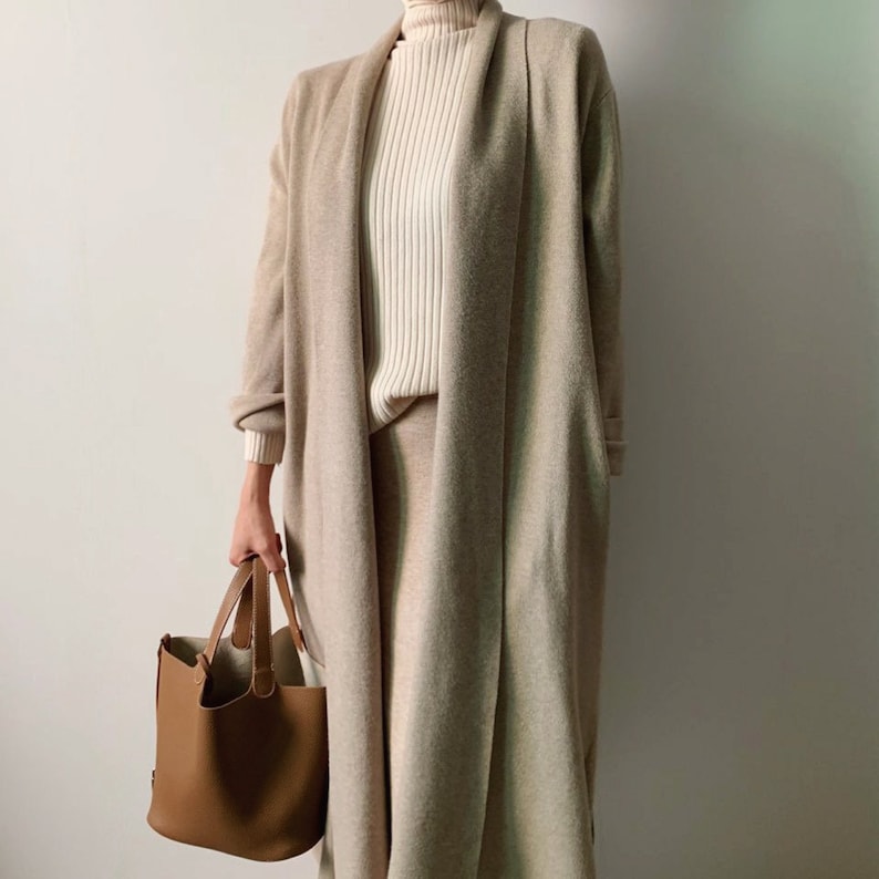 May include: A beige long cardigan and matching skirt are worn over a cream-colored ribbed turtleneck. A brown leather tote bag is held in the hand. The outfit is neutral-toned and minimalist.