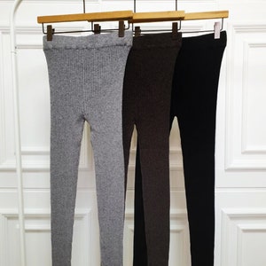 Puede incluir: Tres pares de leggings de punto acanalado en gris, marr&oacute;n y negro, colgados de perchas de madera. Los leggings se exhiben sobre un fondo blanco, mostrando su textura y variaciones de color. La tela parece ser de peso medio.