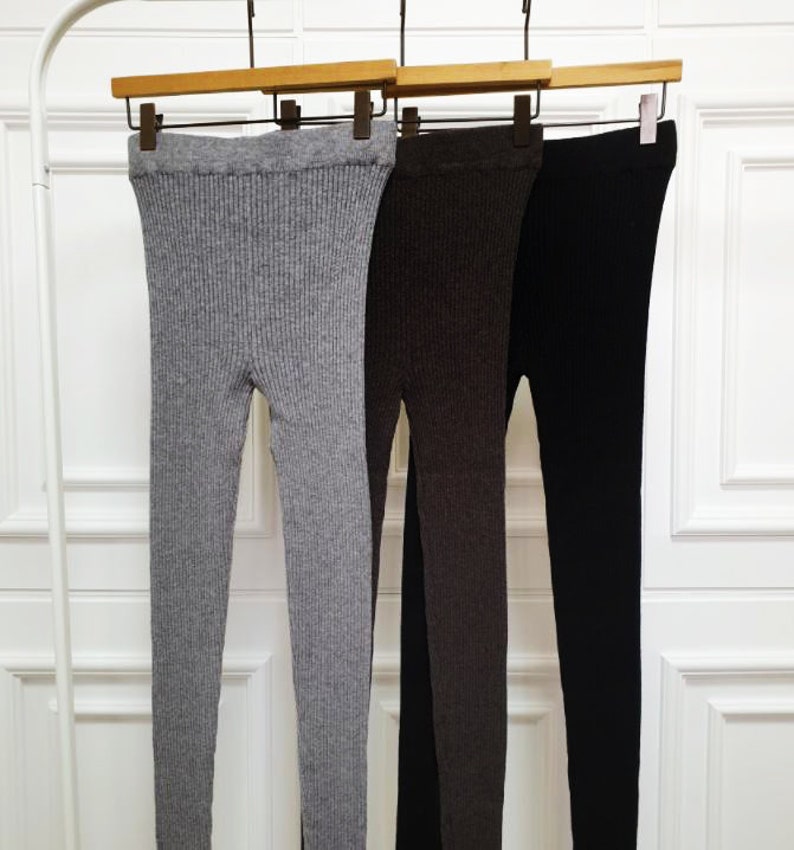 Puede incluir: Tres pares de leggings de punto acanalado en gris, marr&oacute;n y negro, colgados de perchas de madera. Los leggings se exhiben sobre un fondo blanco, mostrando su textura y variaciones de color. La tela parece ser de peso medio.