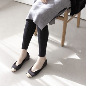 Puede incluir: Leggings negros acanalados y bailarinas negras con punteras doradas. Los leggings se llevan con un su&eacute;ter gris. La escena est&aacute; ambientada en interiores con una silla de madera.