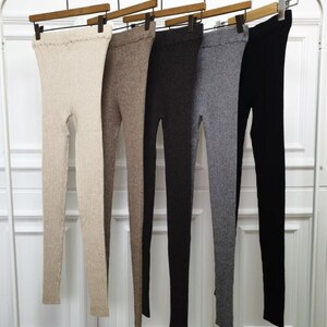 Puede incluir: Cinco pares de leggings de punto acanalado en una variedad de colores neutros cuelgan de perchas de madera. Los colores incluyen beige, marr&oacute;n, gris oscuro, gris claro y negro. Los leggings se muestran sobre un fondo blanco.