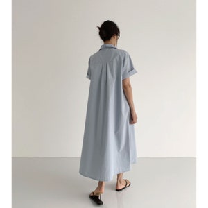 May include: A light blue, short-sleeved shirt dress. The dress has a collared neckline and a button-down front. The dress falls to the mid-calf and has a relaxed fit. The model is wearing black sandals.