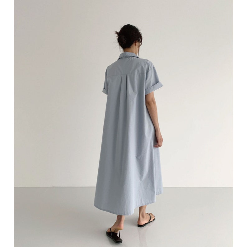 May include: A light blue, short-sleeved shirt dress. The dress has a collared neckline and a button-down front. The dress falls to the mid-calf and has a relaxed fit. The model is wearing black sandals.