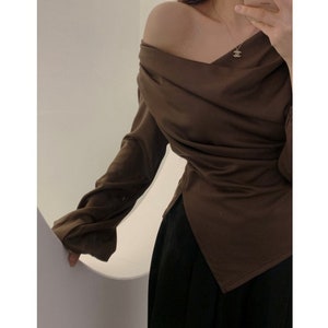 May include: A brown off-the-shoulder top with long sleeves and a draped design. The top has a V-neckline and a split hem. The bottom of the image shows a black skirt.