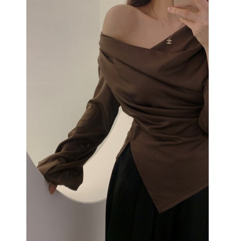 May include: A brown off-the-shoulder top with long sleeves and a draped design. The top has a V-neckline and a split hem. The bottom of the image shows a black skirt.