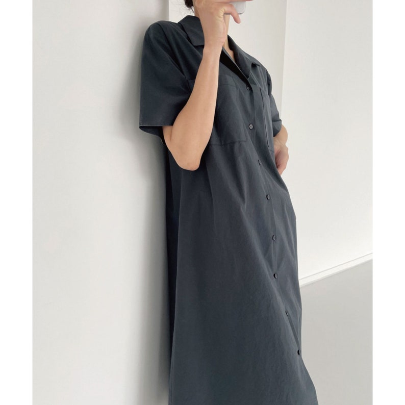 May include: A dark gray, button-down shirt dress with short sleeves and a collared neckline. The dress features a chest pocket and a relaxed fit, suitable for casual wear. The dress is made of a lightweight fabric.