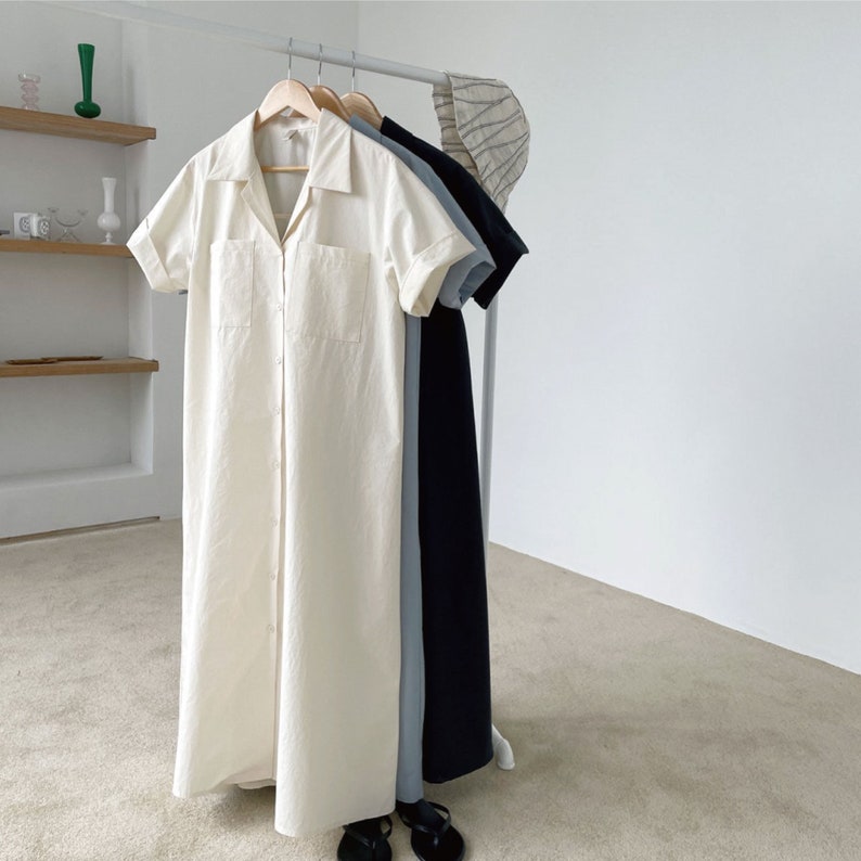 May include: Three button-down dresses in cream, light blue, and navy blue hang on a clothing rack. The cream dress has short sleeves and two front pockets. The light blue and navy dresses also have short sleeves. A striped hat hangs on the right.