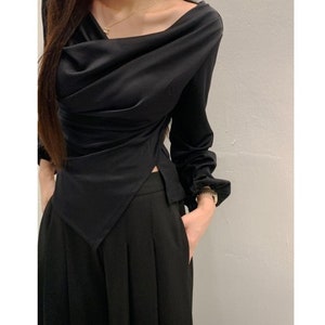 May include: Black long-sleeve top with a draped cowl neckline and a split hem. The top is paired with black pleated pants. The outfit is a sleek, monochromatic ensemble.