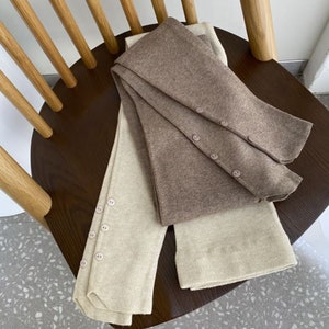 Peut inclure: Jambi&egrave;res en tricot bicolores beige et marron, avec des boutons. Les jambi&egrave;res sont pli&eacute;es sur une chaise en bois, mettant en valeur leur texture et leur design. Les boutons sont de la m&ecirc;me couleur que les jambi&egrave;res.