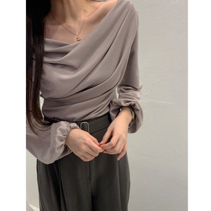 May include: A draped, off-the-shoulder blouse in a muted mauve color. The long sleeves gather at the wrists, and the fabric drapes across the bodice. The blouse is paired with dark gray trousers and a black belt.