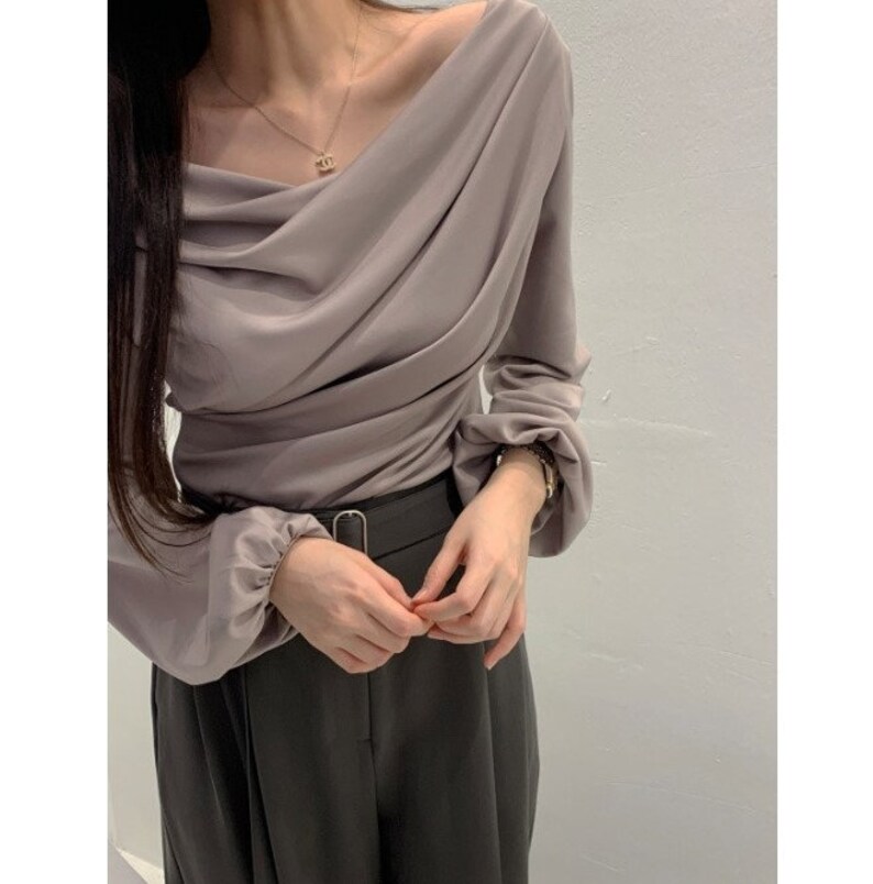 May include: A draped, off-the-shoulder blouse in a muted mauve color. The long sleeves gather at the wrists, and the fabric drapes across the bodice. The blouse is paired with dark gray trousers and a black belt.