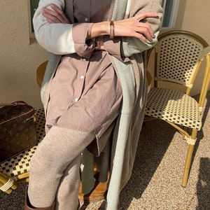May include: A layered outfit featuring a long, color-blocked cardigan in cream, beige, and mint green. The outfit includes a button-down shirt, leggings, and a brown leather bracelet. A designer travel bag and a wicker chair are also visible.