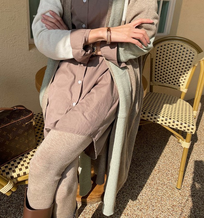 May include: A layered outfit featuring a long, color-blocked cardigan in cream, beige, and mint green. The outfit includes a button-down shirt, leggings, and a brown leather bracelet. A designer travel bag and a wicker chair are also visible.