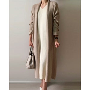 May include: A long, beige cardigan with a shawl collar is worn over a cream-colored, V-neck dress. A white handbag is held in the left hand. The outfit is completed with white shoes and a black watch.