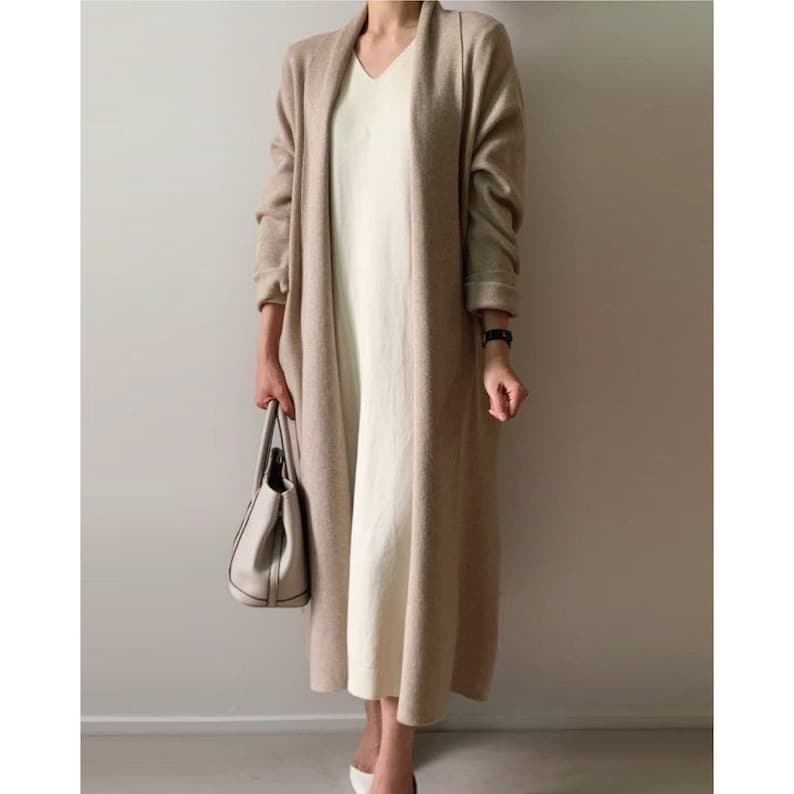 May include: A long, beige cardigan with a shawl collar is worn over a cream-colored, V-neck dress. A white handbag is held in the left hand. The outfit is completed with white shoes and a black watch.