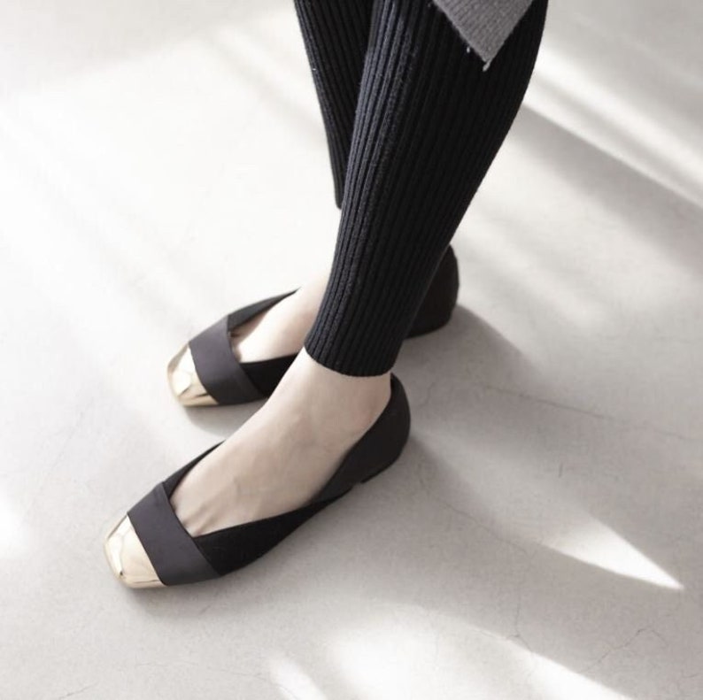 Puede incluir: Bailarinas negras con puntera dorada. Los zapatos tienen punta cuadrada y una correa en la parte superior. La persona lleva leggings negros acanalados.