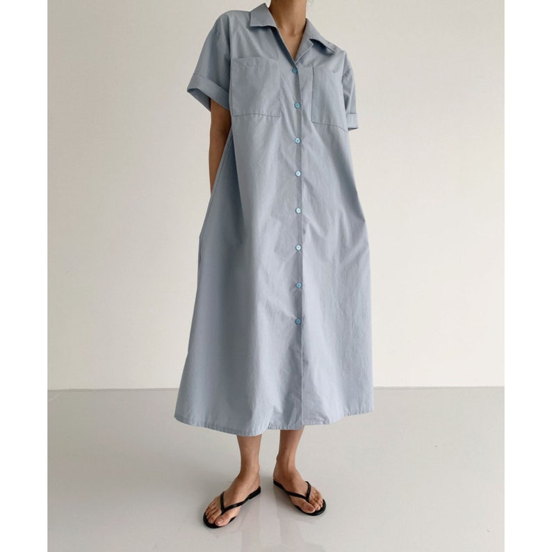 May include: A light blue, button-down shirt dress with short sleeves and two chest pockets. The dress has a collared neckline and a midi length. The buttons and stitching are a matching light blue color. The person is wearing black flip-flops.