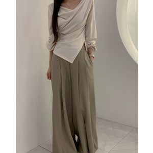May include: A cream-colored long-sleeve top with a draped neckline and asymmetrical hemline. Paired with wide-leg, olive-green trousers. The outfit is a modern, minimalist style, suitable for various occasions.