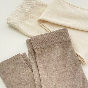 May include: Close-up of three pairs of leggings in neutral tones. One pair is a light beige, another is a creamy off-white, and the third is a muted brown. Each pair has a ribbed waistband and a soft, textured appearance.