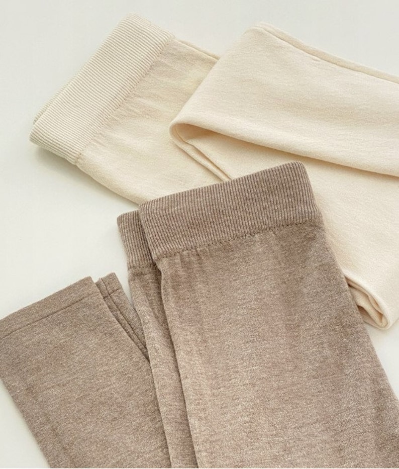 May include: Close-up of three pairs of leggings in neutral tones. One pair is a light beige, another is a creamy off-white, and the third is a muted brown. Each pair has a ribbed waistband and a soft, textured appearance.
