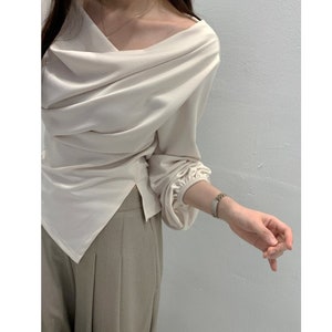 May include: Cream-colored draped blouse with long sleeves and a unique asymmetrical neckline. The blouse has a flowing, elegant design and is paired with light brown pants. The sleeves gather at the wrists, adding a touch of sophistication.