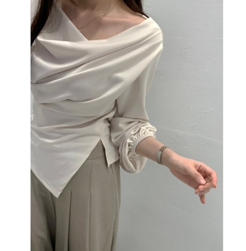 May include: Cream-colored draped blouse with long sleeves and a unique asymmetrical neckline. The blouse has a flowing, elegant design and is paired with light brown pants. The sleeves gather at the wrists, adding a touch of sophistication.