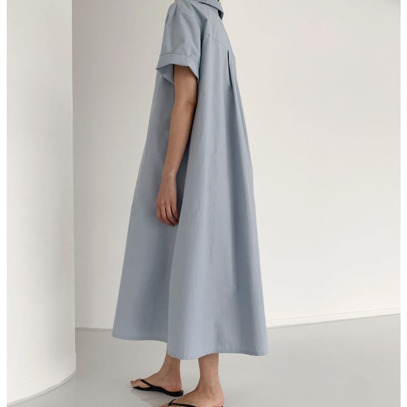 May include: A light blue, short-sleeved dress with a collared neckline and a relaxed fit. The dress has a button-down front and a midi length, suitable for casual wear. The model is wearing black sandals.