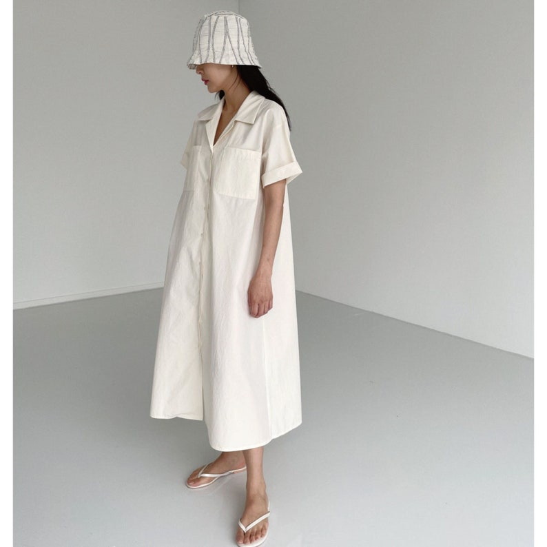 May include: A cream-colored, button-down dress with short sleeves and two front pockets. The dress is paired with a white and gray striped bucket hat and white flip-flops. The dress has a collared neckline and a relaxed fit.