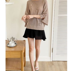 May include: A woman's outfit featuring a tan, loose-fitting top with wide sleeves and a black, pleated mini skirt. The outfit is paired with white sandals. A small table with a tea set is in the background.