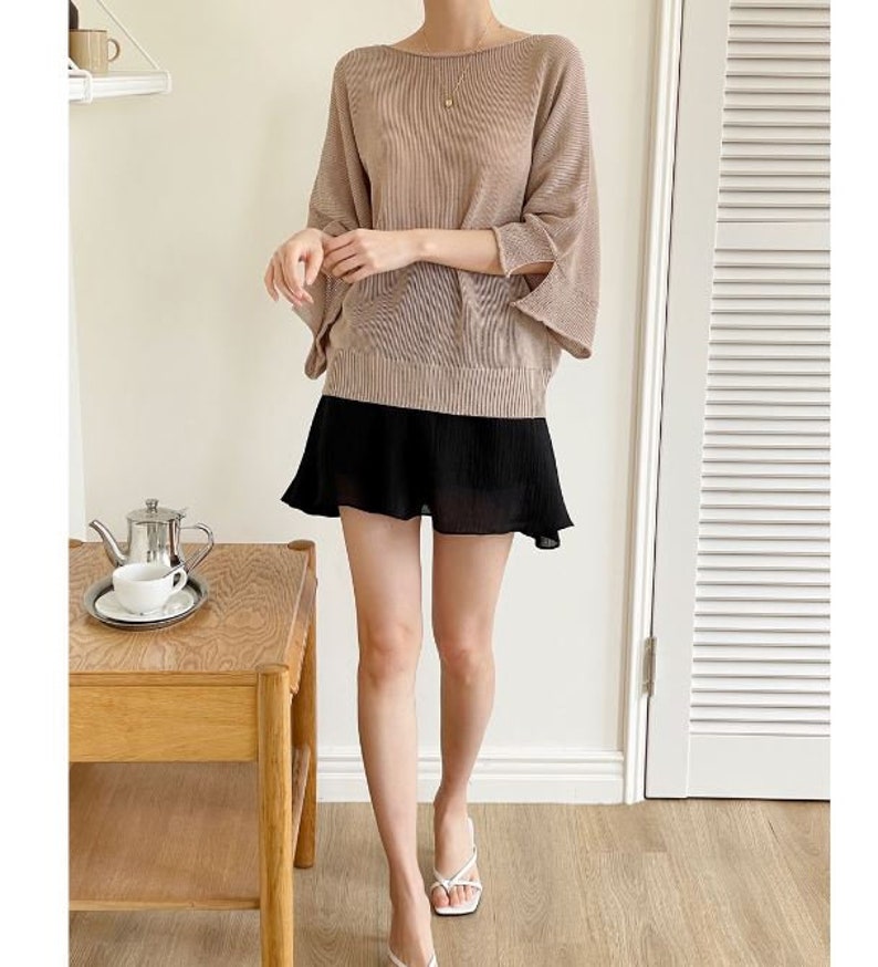 May include: A woman's outfit featuring a tan, loose-fitting top with wide sleeves and a black, pleated mini skirt. The outfit is paired with white sandals. A small table with a tea set is in the background.