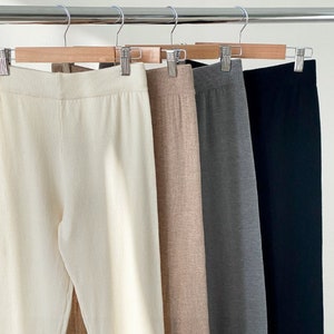 May include: Four pairs of knit pants in cream, tan, gray, and black hang on a wooden clothes rack. The pants have a ribbed waistband and a straight leg design. The fabric appears to be a soft, lightweight knit.