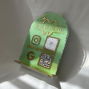 May include: A green and gold acrylic sign with the text "Dent Bomont" and a stylized tooth graphic. The sign also includes social media icons for Instagram and Google, as well as a QR code. The sign is on a white background.