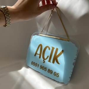 May include: A light blue acrylic sign with the word "ACIK" in gold lettering and the phone number "0551 905 89 05" printed below. The sign is hanging from a gold ribbon.