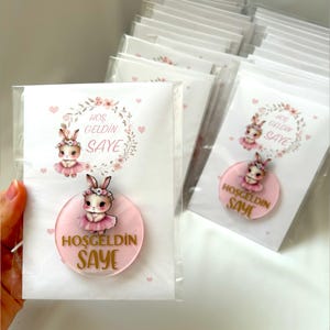Könnte beinhalten: Ein Satz rosa und goldener Gastgeschenketiketten mit einer Hasenillustration und dem Text "Hos geldin Saye", was "Willkommen Saye" bedeutet.