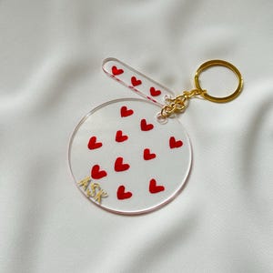 Puede incluir: Un llavero de acrílico transparente con un dije circular con corazones rojos y la palabra "ASK" en letras doradas. El llavero también incluye un dije rectangular más pequeño con corazones rojos.