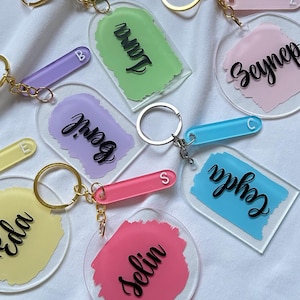 May include: Six colourful acrylic keychains with different names written on them. Each keychain has a small, clear acrylic tag with a letter on it. The keychains are on a white fabric background.
