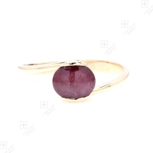 May include: A gold ring with a single, round, dark pink gemstone set in the center. The ring band is thin and wraps around the finger in a simple, elegant design.