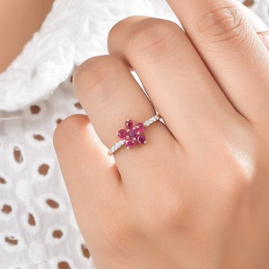 Può includere: Un anello d'oro con un gruppo di pietre preziose rosse incastonate a forma di fiore. L'anello è circondato da pietre preziose bianche più piccole.