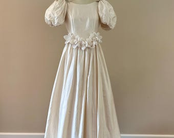 May include: A floor-length cream-colored dress featuring short puffed sleeves and a wide waistband adorned with a row of fabric flowers. The garment has a round neckline and a full, flowing skirt, displayed on a mannequin against a plain wall.