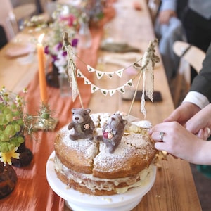May include: A two-tiered sponge cake topped with two needle-felted brown bears and a miniature bunting banner that reads JUST MARRIED. The bears are decorated with small floral accents, and the cake is dusted with powdered sugar on top.