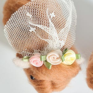 May include: A needle-felted brown animal figure adorned with a white mesh veil and a floral crown featuring small pink, cream, and yellow fabric roses with green ribbon leaves. The figure has a soft, fuzzy texture and a dark, round eye visible.