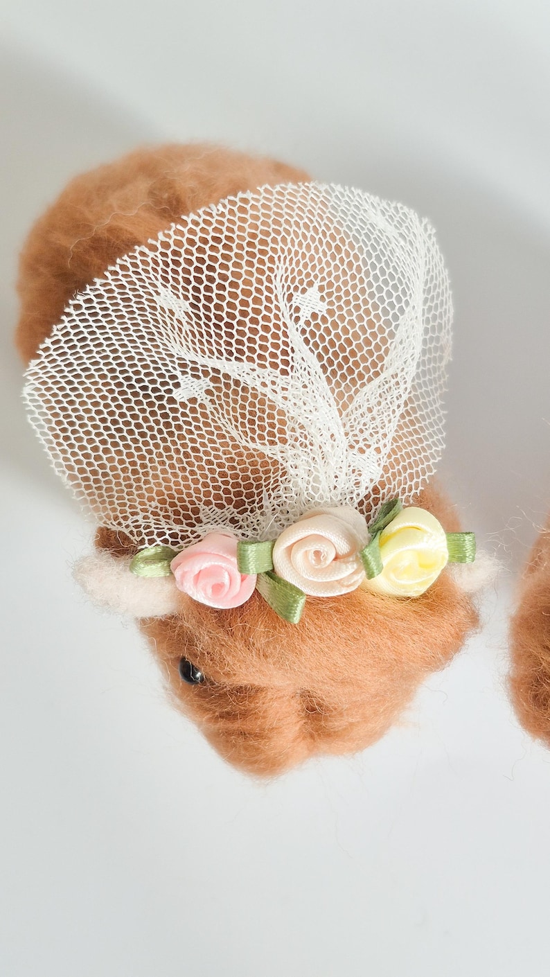 May include: A needle-felted brown animal figure adorned with a white mesh veil and a floral crown featuring small pink, cream, and yellow fabric roses with green ribbon leaves. The figure has a soft, fuzzy texture and a dark, round eye visible.
