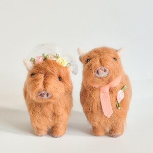 May include: Two needle-felted brown Highland cow figures. The left cow wears a white mesh veil with small pink and yellow fabric flowers. The right cow wears a light pink necktie and a single pink fabric flower pinned to its chest.