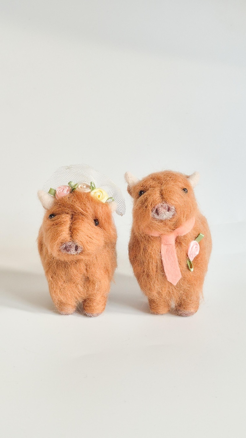 May include: Two needle-felted brown Highland cow figures. The left cow wears a white mesh veil with small pink and yellow fabric flowers. The right cow wears a light pink necktie and a single pink fabric flower pinned to its chest.