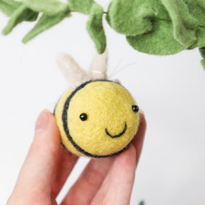 May include: A yellow felt bee with black stripes and a smiling face. The bee has a white felt antenna and is held in a hand.
