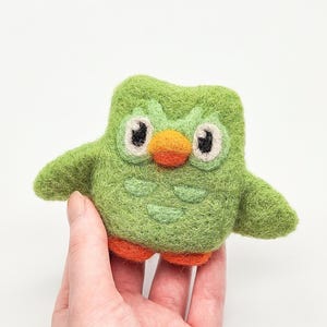 May include: A handmade green felt owl figurine with orange feet and beak, and large white-rimmed eyes. The owl has small wings and is held in a hand against a white background. The owl is approximately 3 inches tall.